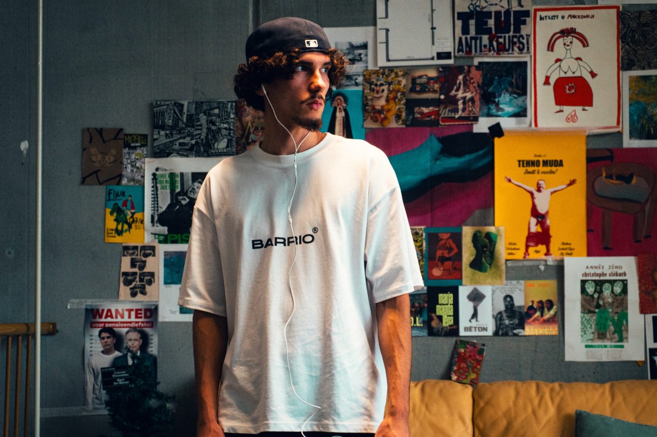 Model met krullen, een achterstevoren pet en witte oortjes in een wit Barrio-T-shirt, voor een muur vol kleurrijke posters