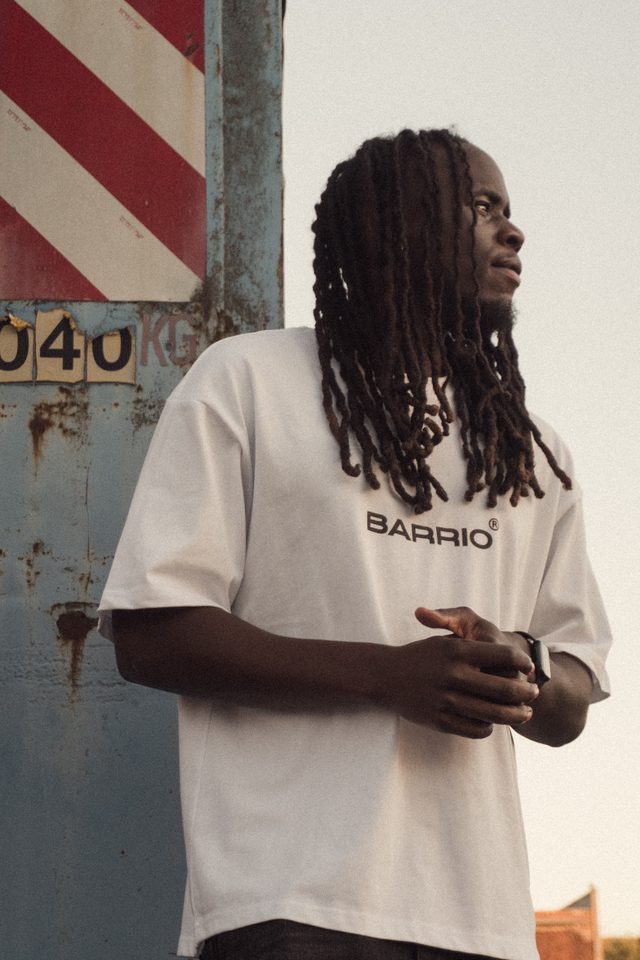 Model met lange dreadlocks in een wit Barrio-T-shirt tegen een roestige blauwe container met een rood-wit gestreept bord