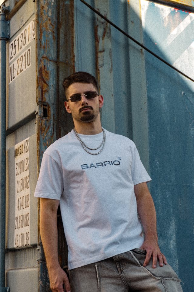 Model met een smalle zonnebril en zilveren kettingen in een wit Barrio-T-shirt, leunend tegen een blauwe zeecontainer met gewichtslabels