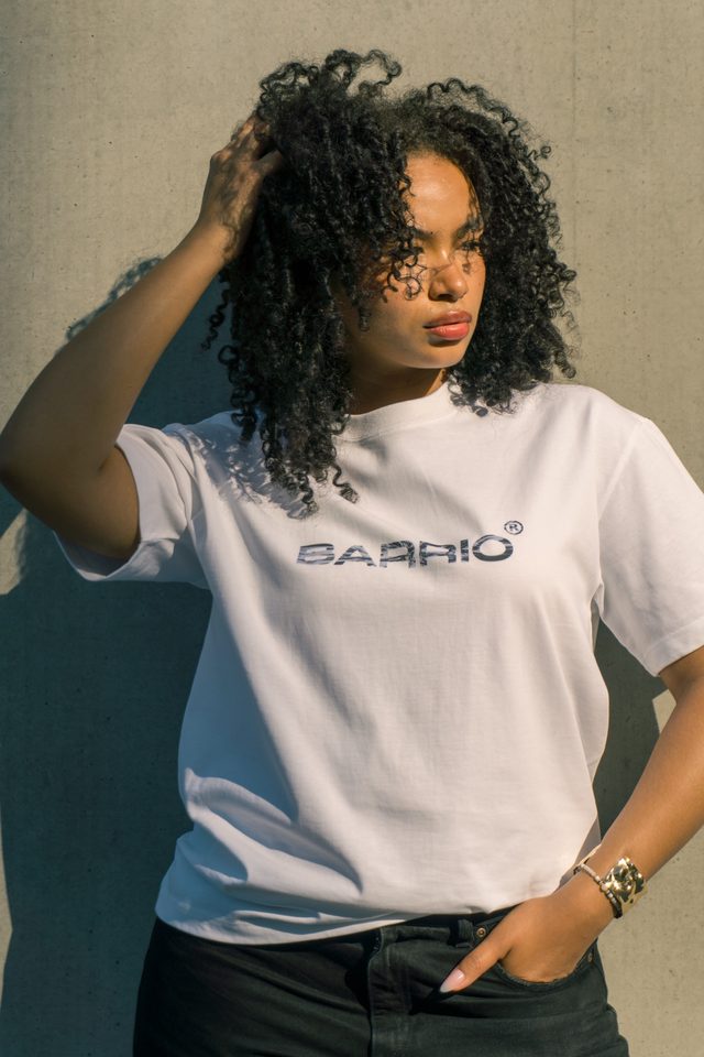 Model met volle krullen en een hand in het haar, in een wit Barrio-T-shirt tegen een betonnen muur in fel zonlicht