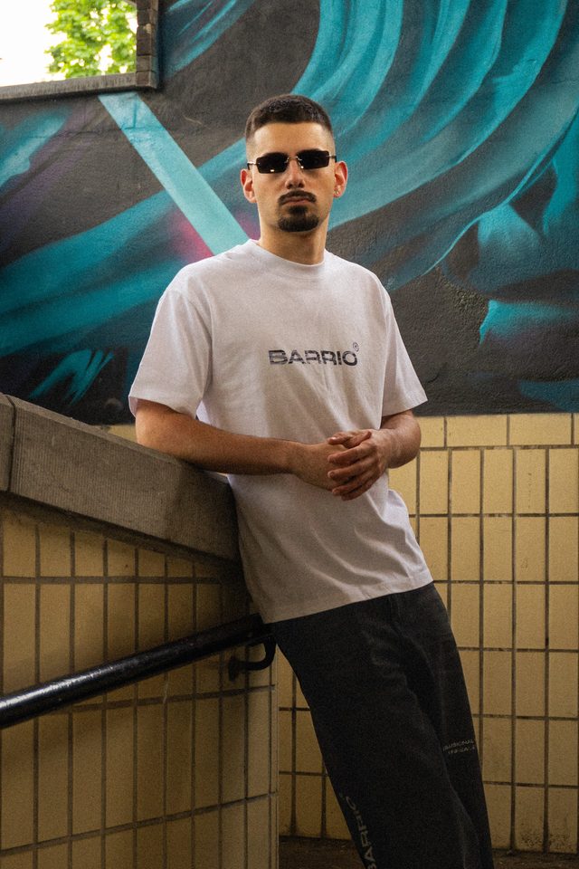Model met zonnebril in een wit Barrio-T-shirt, leunend op een trapmuur in een betegelde onderdoorgang met een turquoise muurschildering