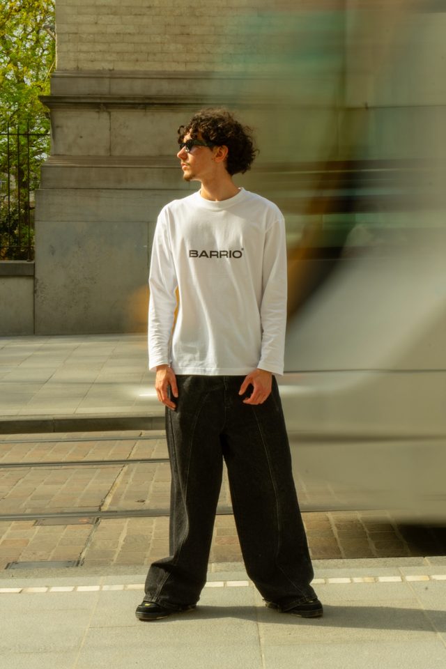 Model met krullen en zonnebril in een witte Barrio-longsleeve en een wijde zwarte jeans op een perron, terwijl een wazige tram voorbijrijdt