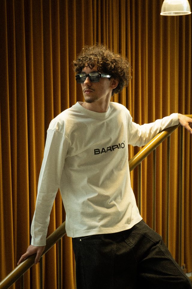 Model met krullen en zonnebril in een witte Barrio-longsleeve, leunend op een goudkleurige trapleuning voor een okergeel gordijn